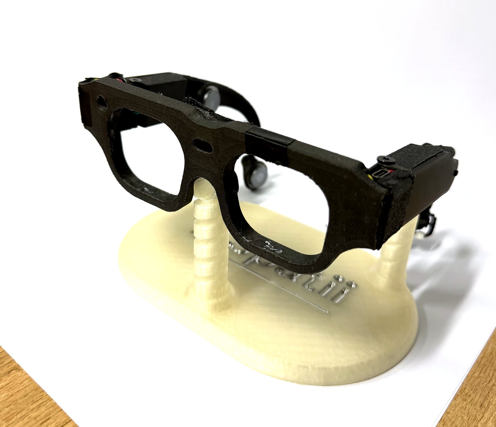 Photograph of the revision 4 prototype on a 3D-printed Spatii display stand: a matte black spectacle frame with bone-conduction transducer arms curving behind each temple.
